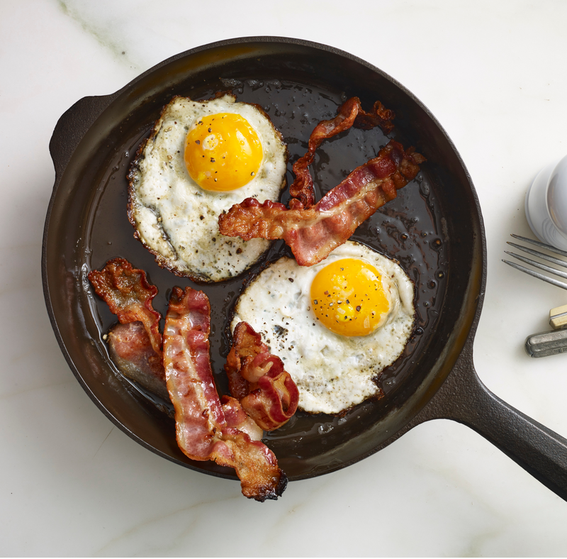 Crispy bacon and sunny-side-up eggs cooking in a smooth cast iron skillet on a white countertop