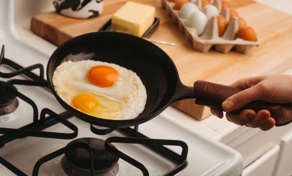 Person holding a No.5 Field Cast Iron Skillet with two fried eggs over a gas flame, eggs and butter behind
